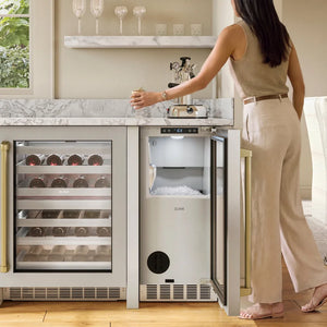 A woman stands beside a built-in undercounter ZLINE Autograph Edition 15 in. Touchstone Nugget Ice Maker with Drain Pump and Solid Stainless Steel Door with Champagne Bronze Handle (INGDZ-ST-15-CB) in a bright kitchen or bar area.
