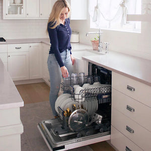 A woman stands in a bright white kitchen next to an open ZLINE Autograph Edition 24 in. Tallac Series 3rd Rack Dishwasher with Auto Door Open Drying, 51dBa, Panel Ready with Matte Black Handle (DWVZ-24-MB), loading plates and glassware into the lower rack.
