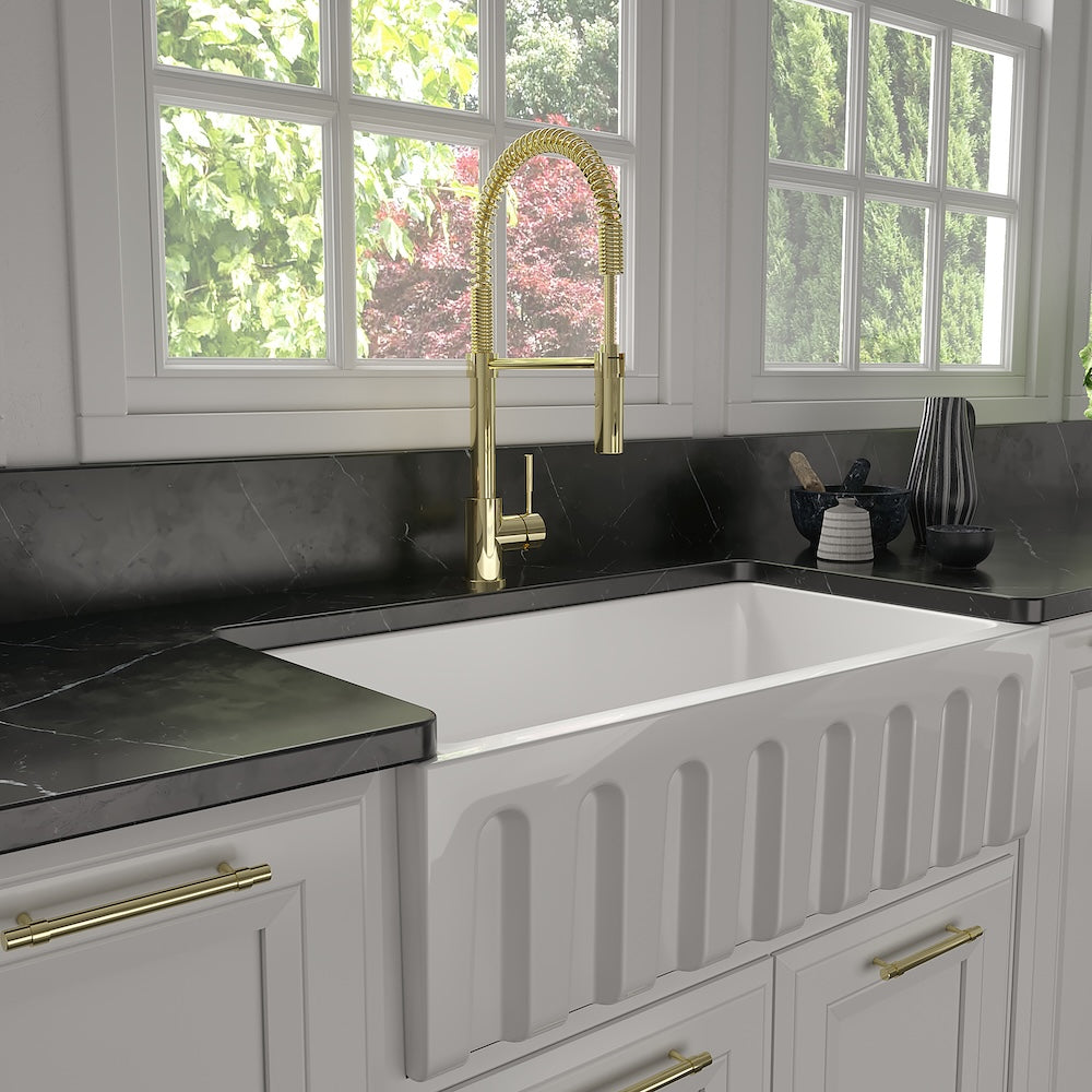 ZLINE Sierra Pull Down Spring Single Handle Kitchen Faucet in Polished Gold (SRA-KF-PG) in a kitchen with a white farmhouse sink, black countertop, and gold accents.