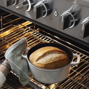 ZLINE 36 in. 5.2 cu. ft. Marino Gas Range with Convection Gas Oven in Stainless Steel with 6 Brass Burners (MGR-BR-36) close-up, a cast-iron Dutch oven with white handles sits on an oven rack inside an oven, with a loaf of bread baking inside. A hand holding a gray cloth is visible on the left.