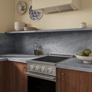 A modern kitchen scene featuring a ZLINE 24 in. 3.2 cu. ft. Marino Induction Range with 3 Element Cooktop and Electric Convection Oven in Stainless Steel (MINR24), set into warm wood cabinets with a gray stone backsplash and matching countertop. Decorative ceramic plates and a range hood are mounted on the cream-colored wall above.