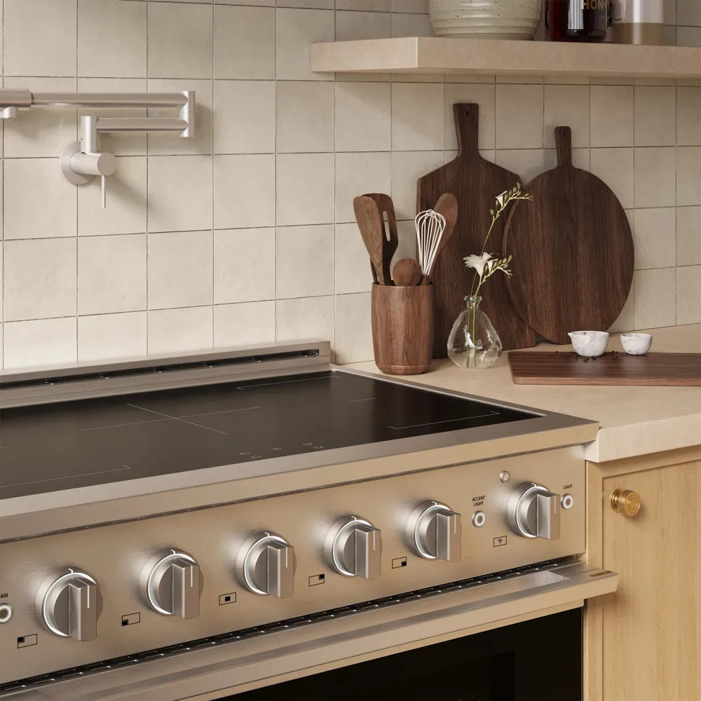A ZLINE 36 in. 5.2 cu. ft. Marino Induction Range with 5 Element Cooktop and Electric Convection Oven in Stainless Steel (MINR36) set into a light wood kitchen cabinet. The background features beige square tiles, a wooden shelf, and dark wood cutting boards on the countertop.