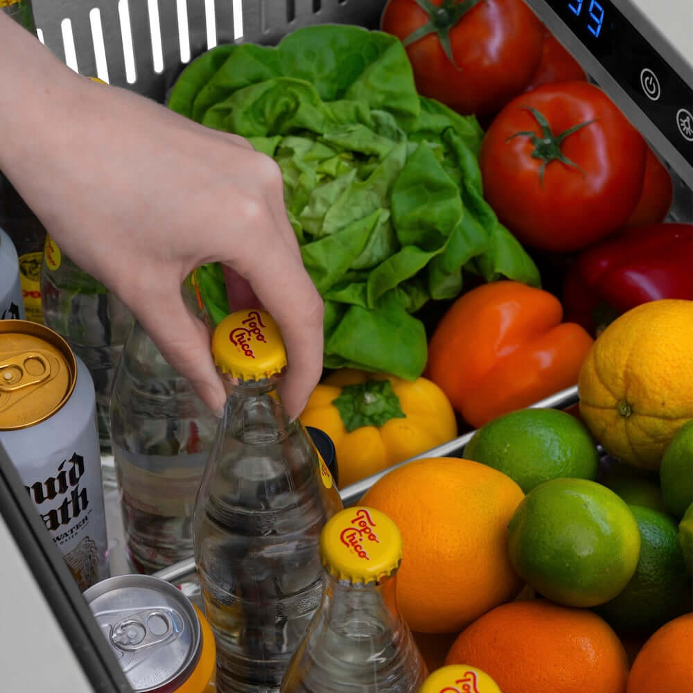 Hand reaching for a bottle amidst fruits and vegetables in a ZLINE 24 in. Touchstone 168 Can Outdoor-Rated Dual Refrigerator Drawer with Hand Hammered Copper Doors (RDSO-HH-24).